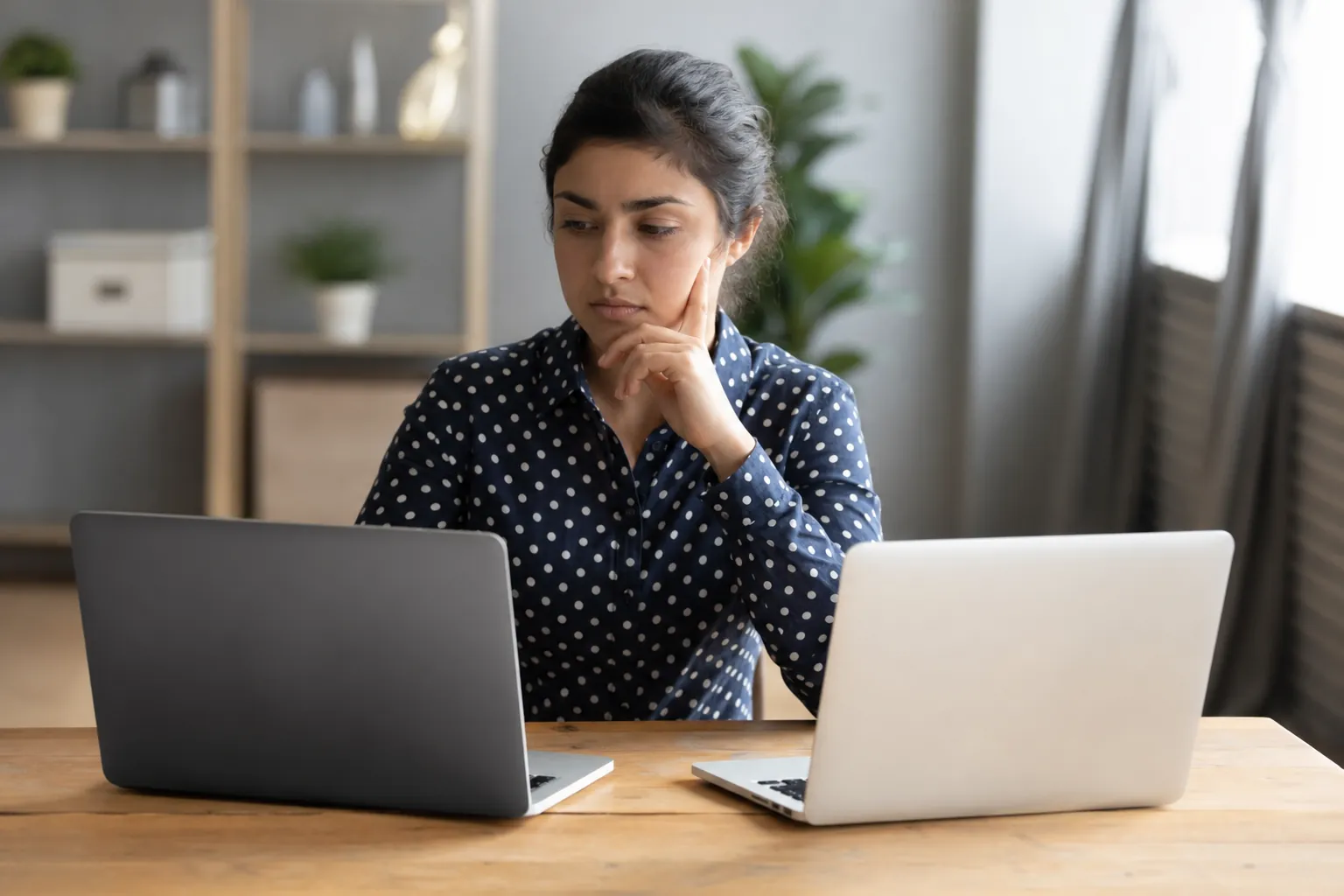 A woman in a navy polka-dot blouse rests her hand against her face while looking at two laptops on a wooden table.