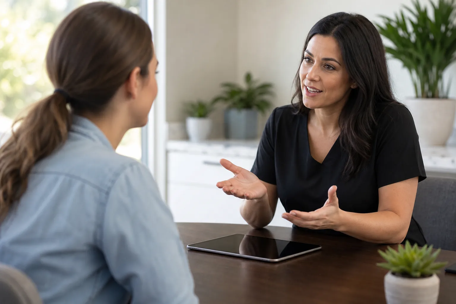 A woman in black scrubs gestures while speaking with a seated woman across a desk with a tablet and potted plant.