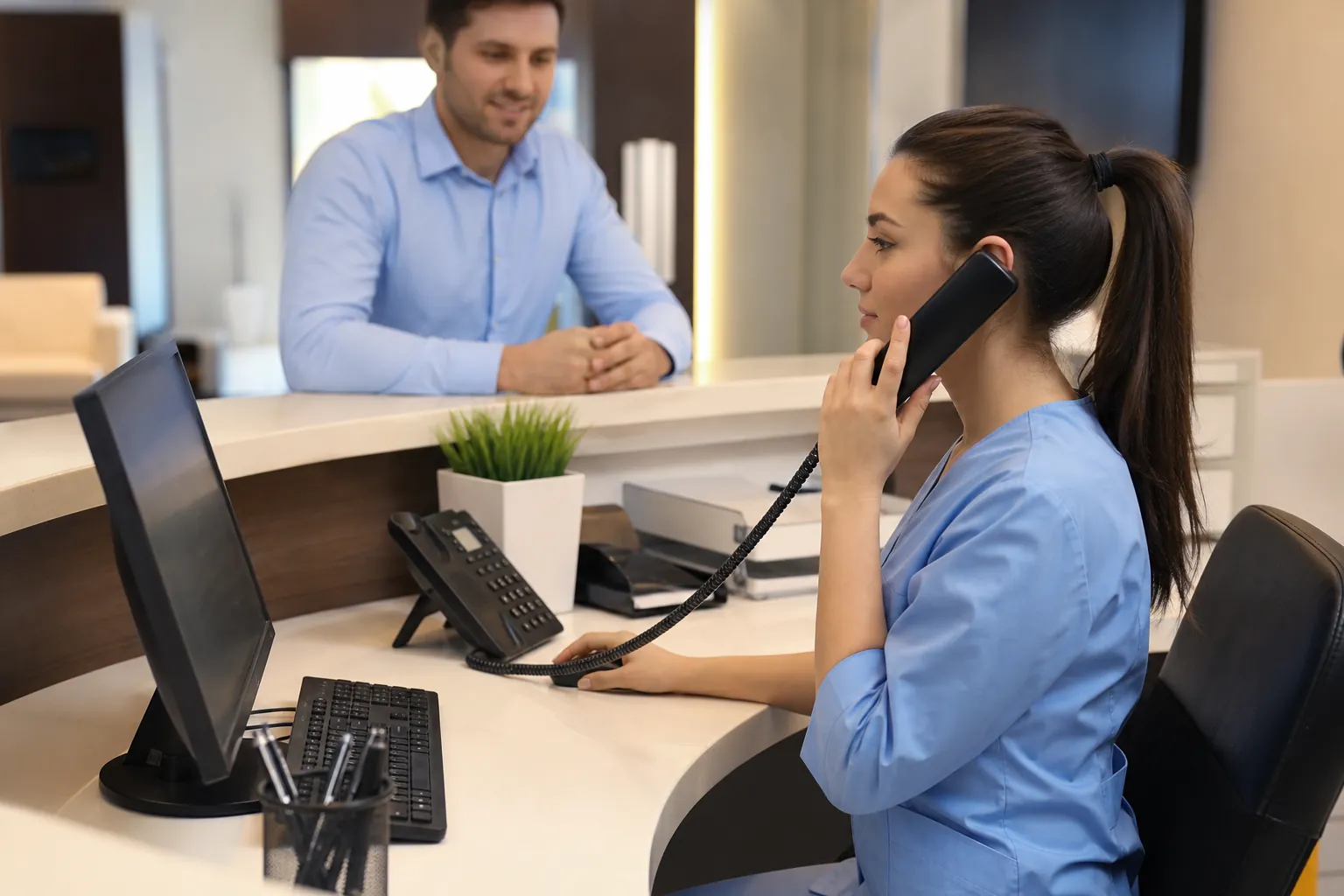 Receptionist in blue scrubs answers a desk phone beside a computer while a man waits across the reception counter.