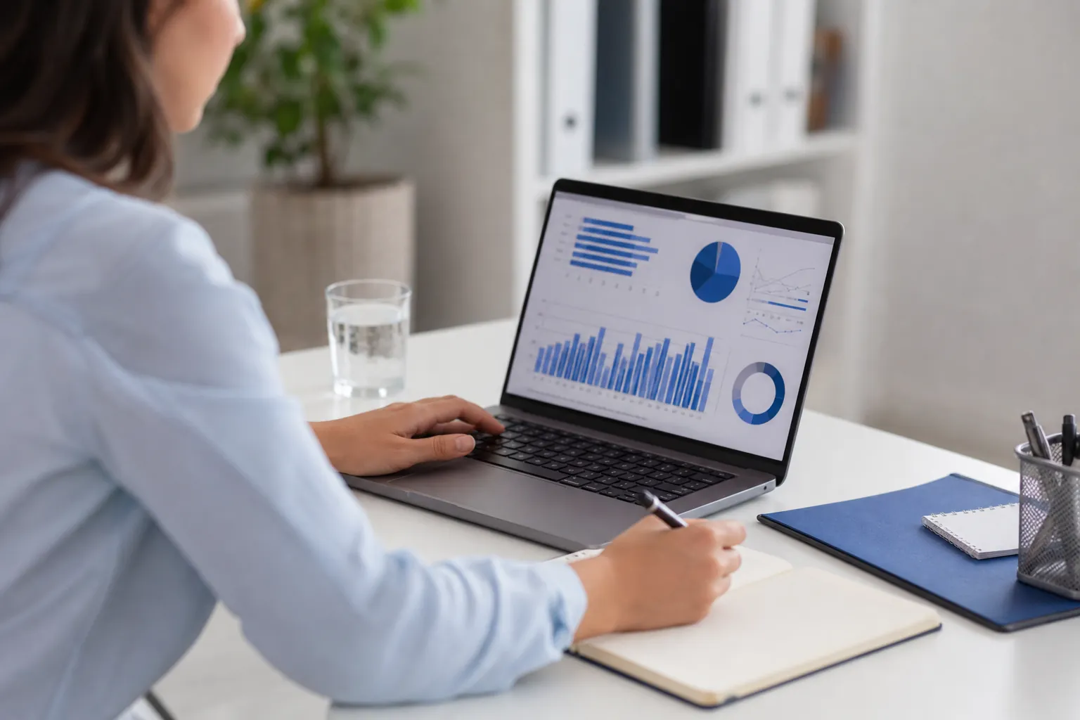 Woman reviewing blue charts on a laptop while holding a pen over an open notebook at a white desk.