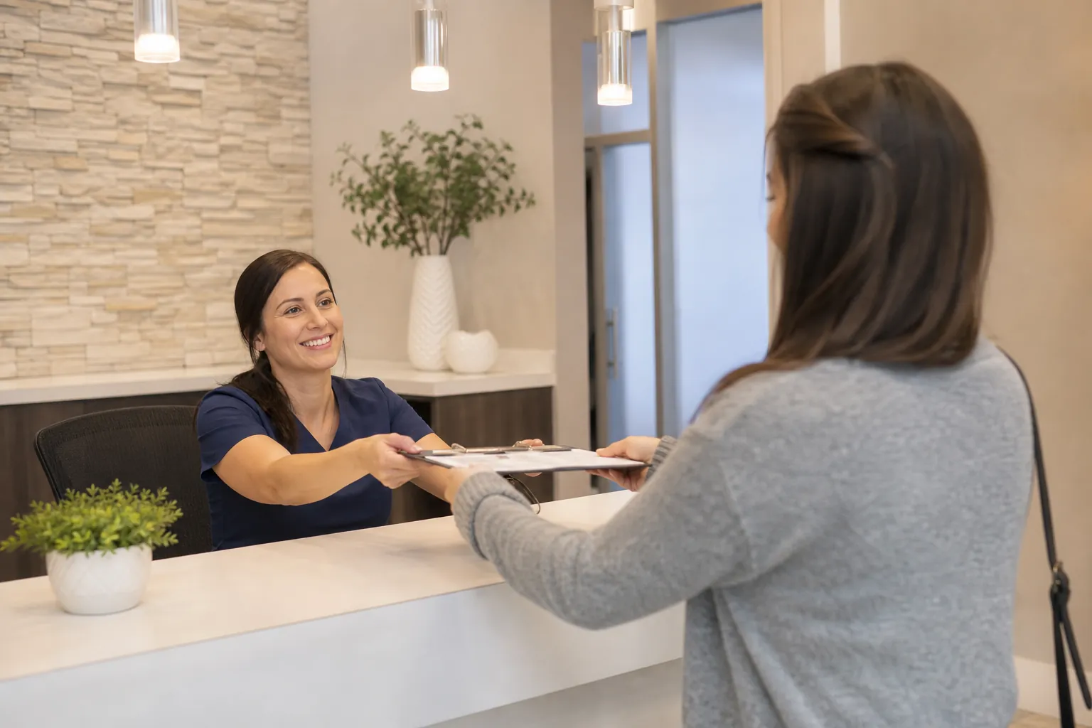 A smiling receptionist in navy scrubs hands a clipboard to a woman across a white reception counter.