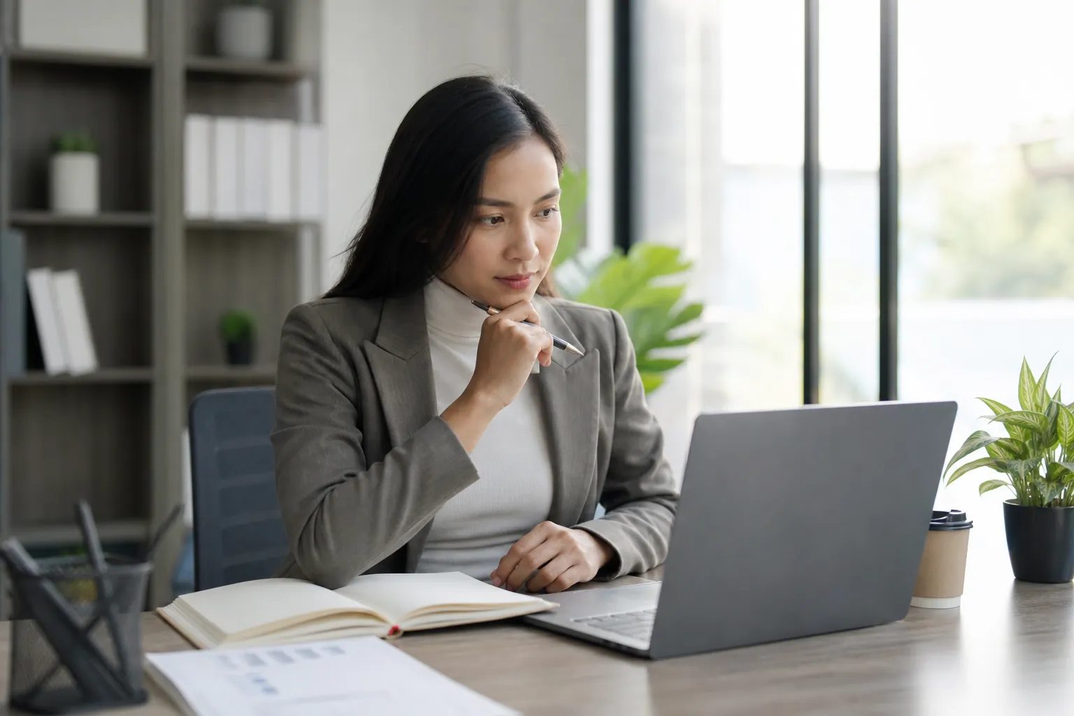 Businesswoman Researching Questions Before Publishing Answers Businesswoman holding a pen while researching questions on a laptop beside an open notebook in a bright office.