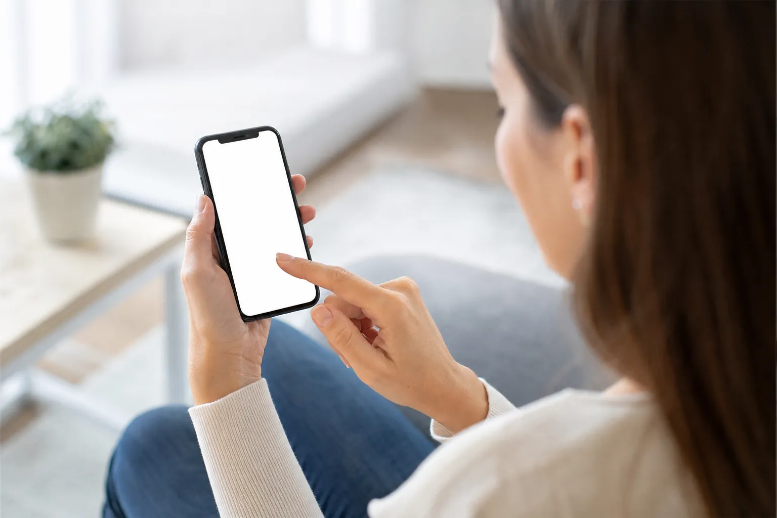 A seated woman holds a smartphone with a blank white screen and touches it with her index finger, viewed over her shoulder.