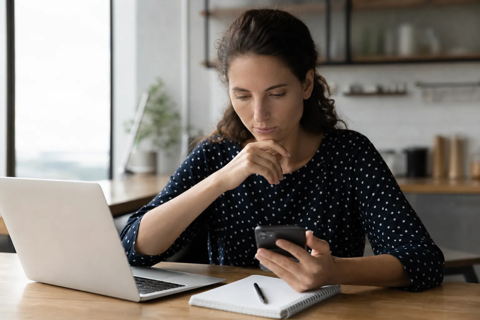 A woman looks at a smartphone while seated at a wooden table with an open laptop, spiral notebook, and pen.
