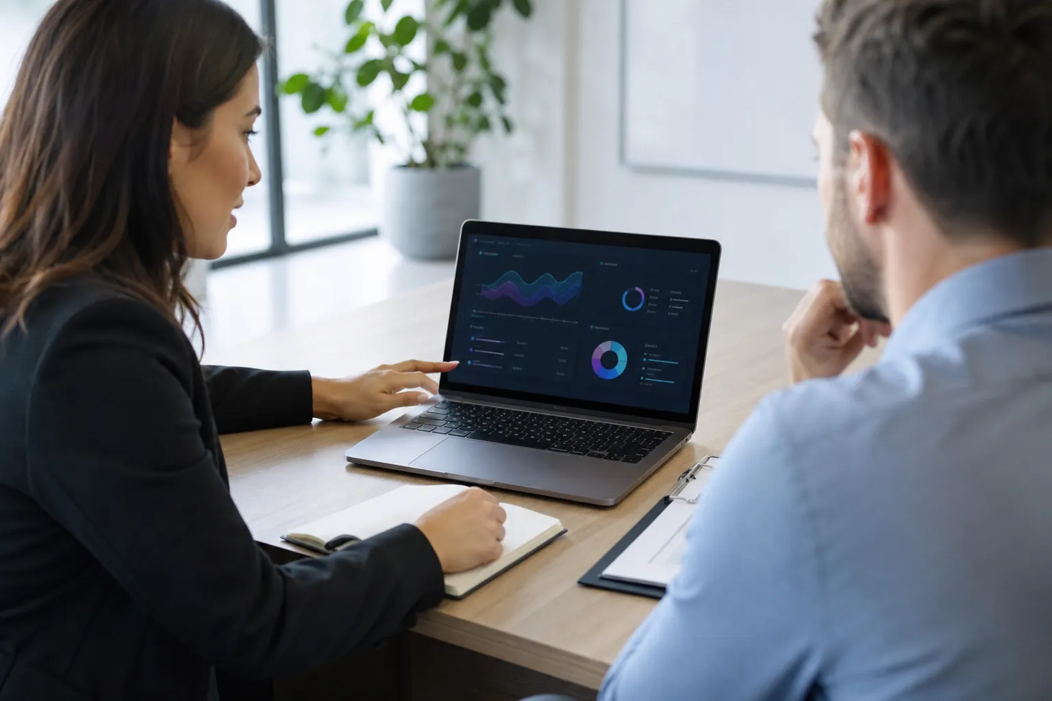 Consultant points to analytics charts on a laptop while meeting with a client across an office table.