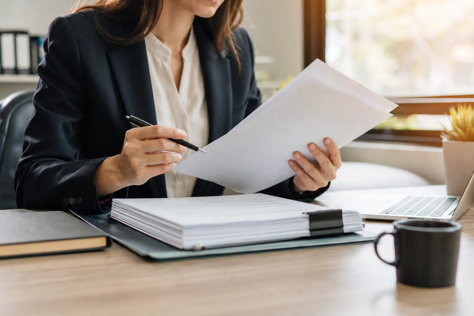 A woman in a dark blazer reviews papers with a pen at a desk holding a thick document stack, laptop, notebook, and black mug.