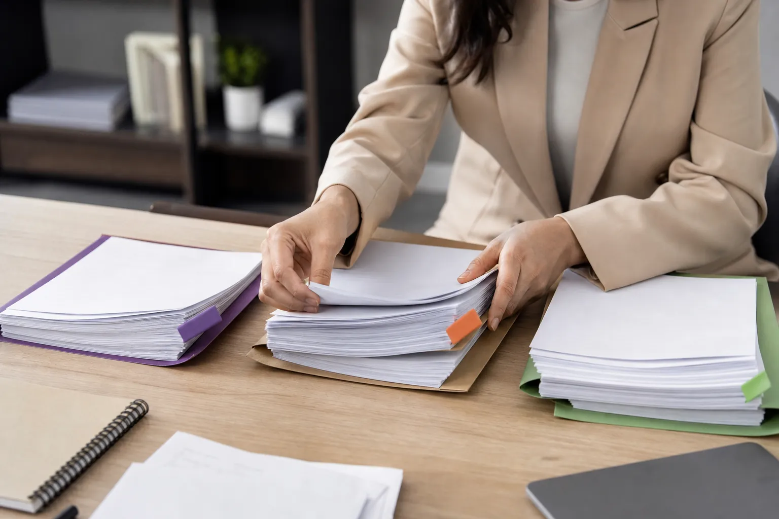 A person in a beige blazer sorts stacks of white papers into purple, brown, and green folders on a wooden desk.