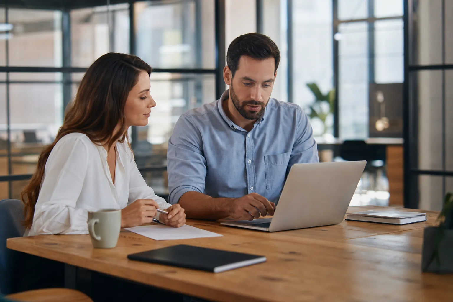 A woman holding a pen and a man using a laptop sit at a wooden office table with paper, notebooks and a mug.