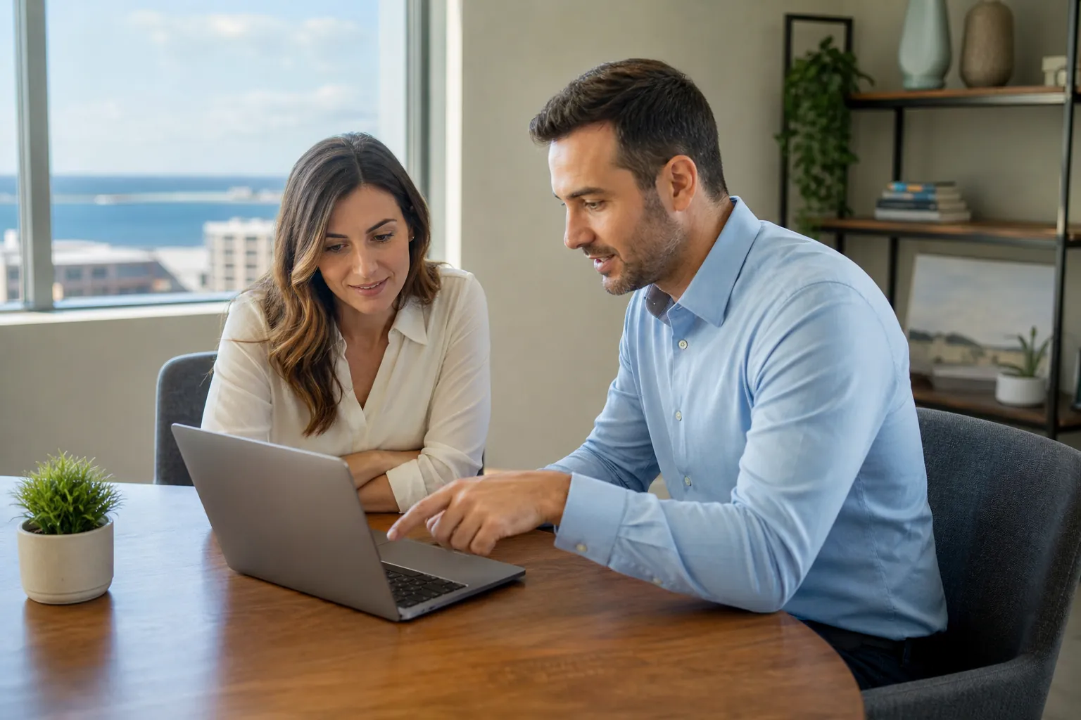 A man points at a laptop while a woman watches across a wooden table in an office overlooking the water.
