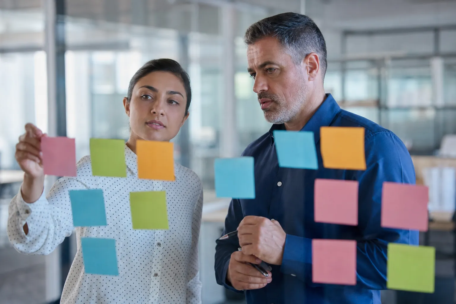 Two people examine blank pink, green, yellow, and blue sticky notes on glass, with the woman reaching toward a pink note.