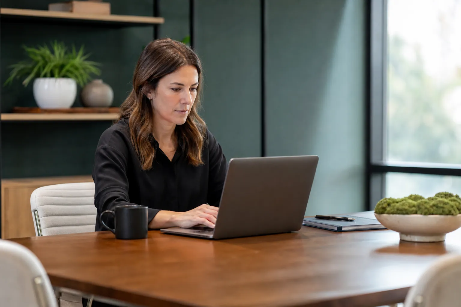 Woman working on a laptop at a wooden table with a black mug, notebook and pen beside a large window.