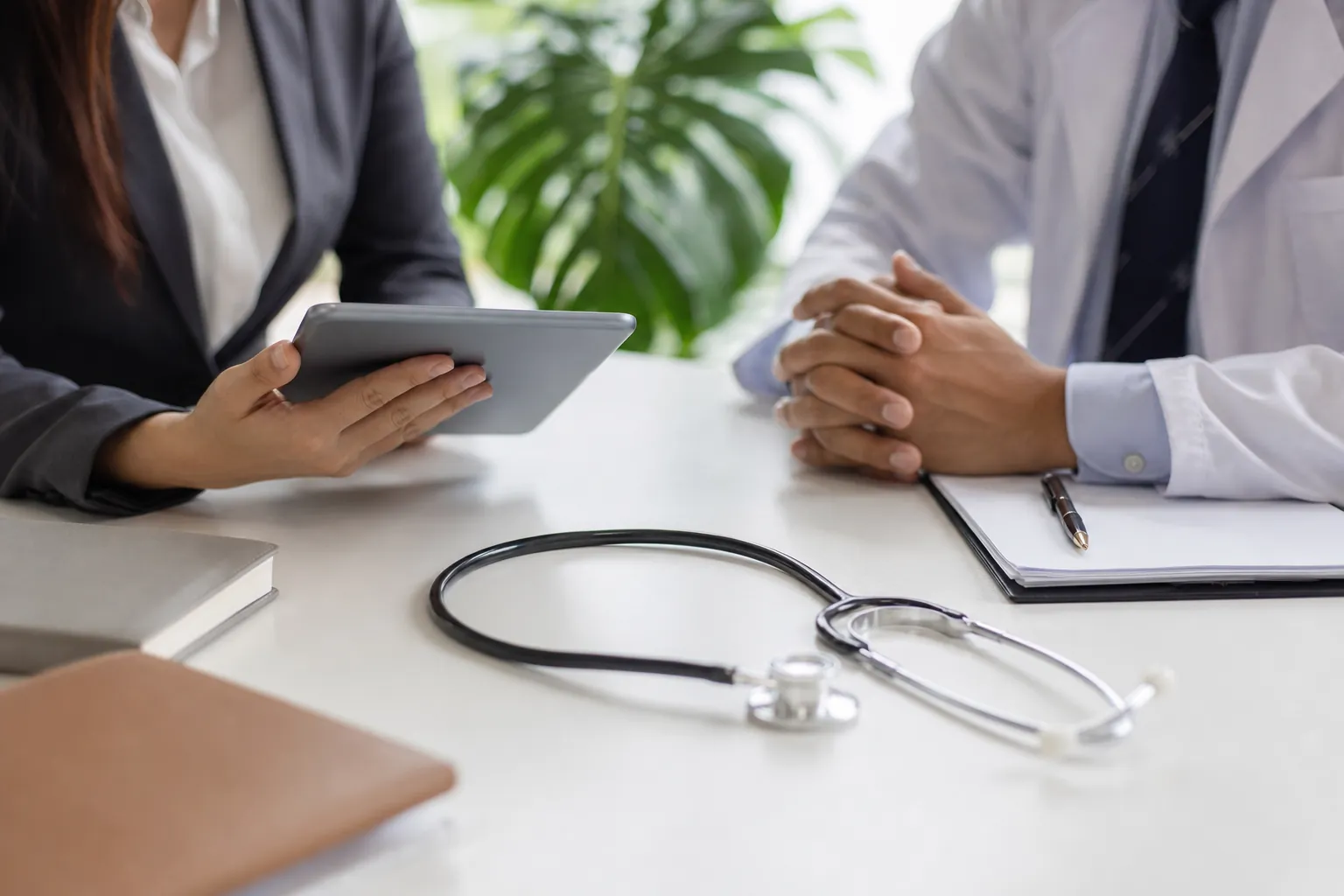 Businesswoman and Doctor Meeting at a Desk A businesswoman holds a tablet across from a doctor in a white coat, with a stethoscope and clipboard on the desk.