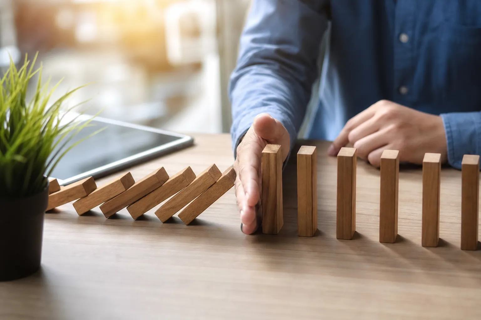 A person in a blue shirt stops falling wooden domino blocks with one hand on a desk beside a tablet and potted plant.