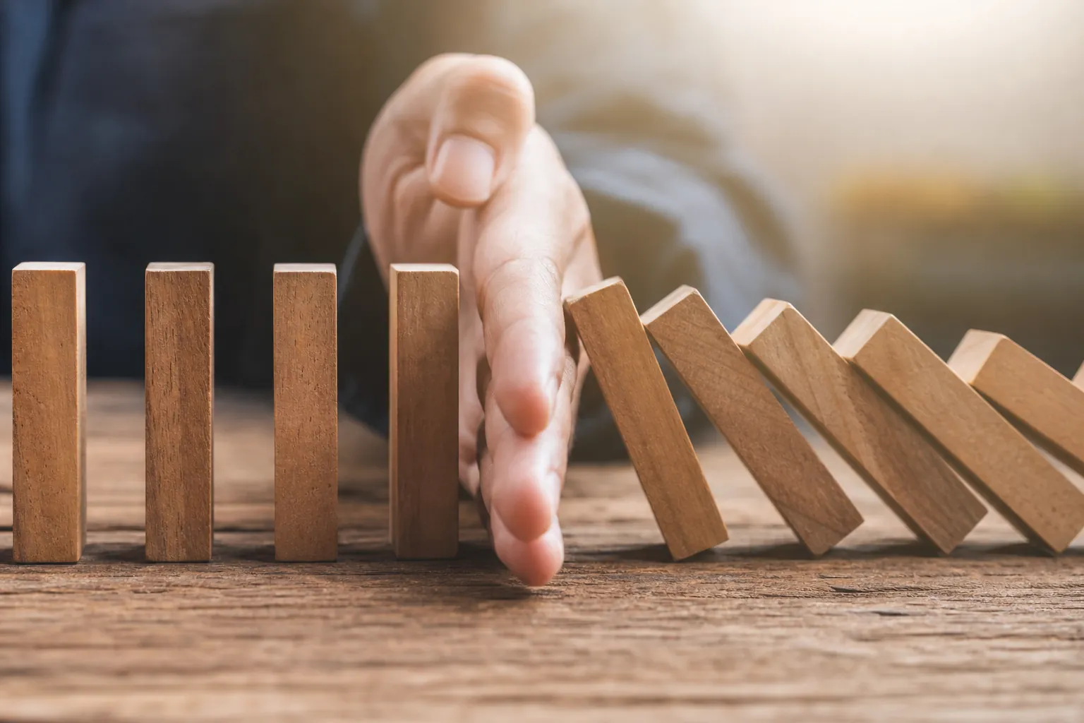 A person's hand stops a row of falling wooden dominoes, protecting upright blocks on a rustic wooden table.